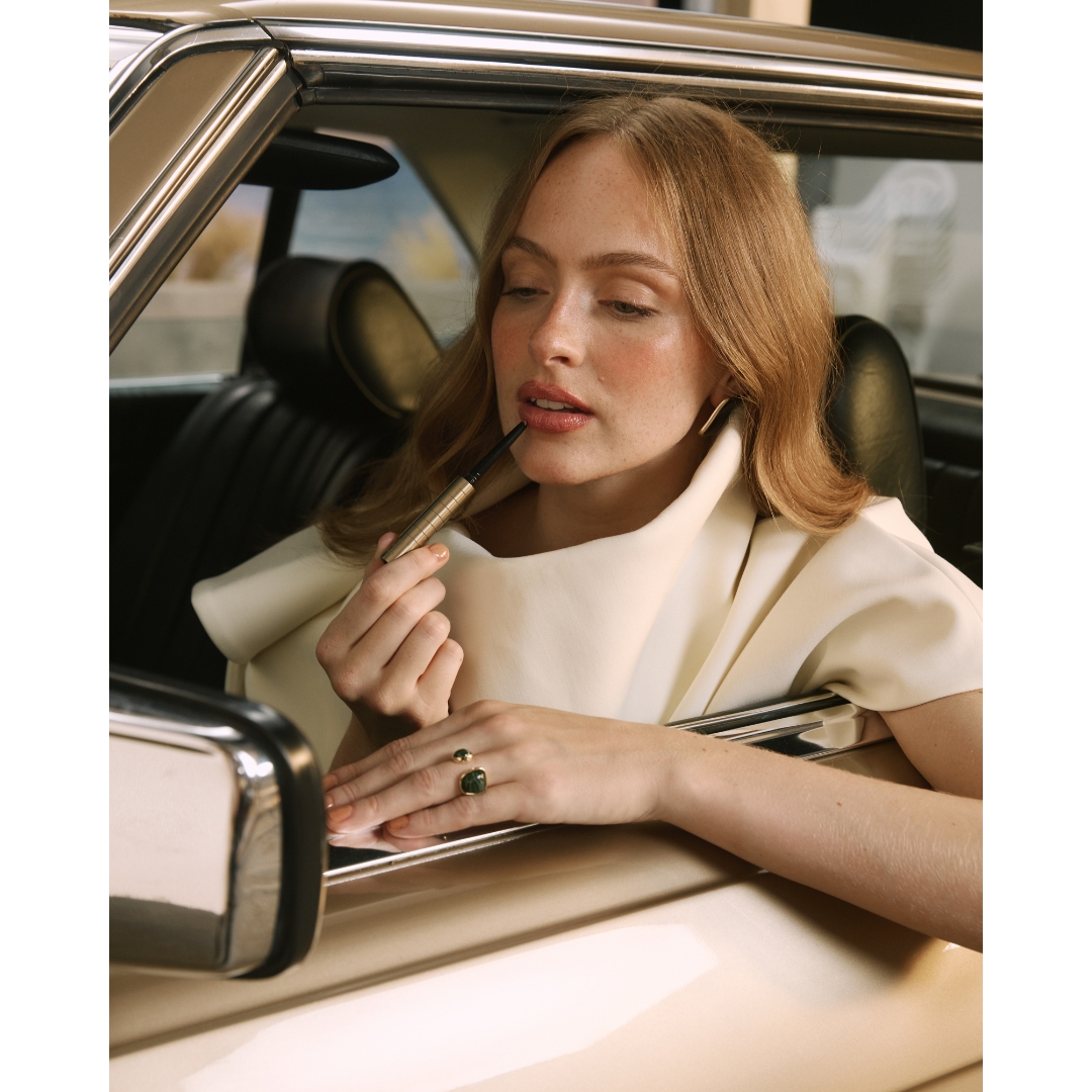 A woman with auburn hair and freckles leaning out of a cream vintage car window, applying lip liner with a gold Karen Betts KB Pen.