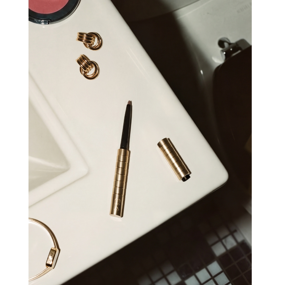 A gold Karen Betts KB Pen with its cap removed, lying on a white sink beside gold knot earrings, a gold bangle, and a pink blush compact.