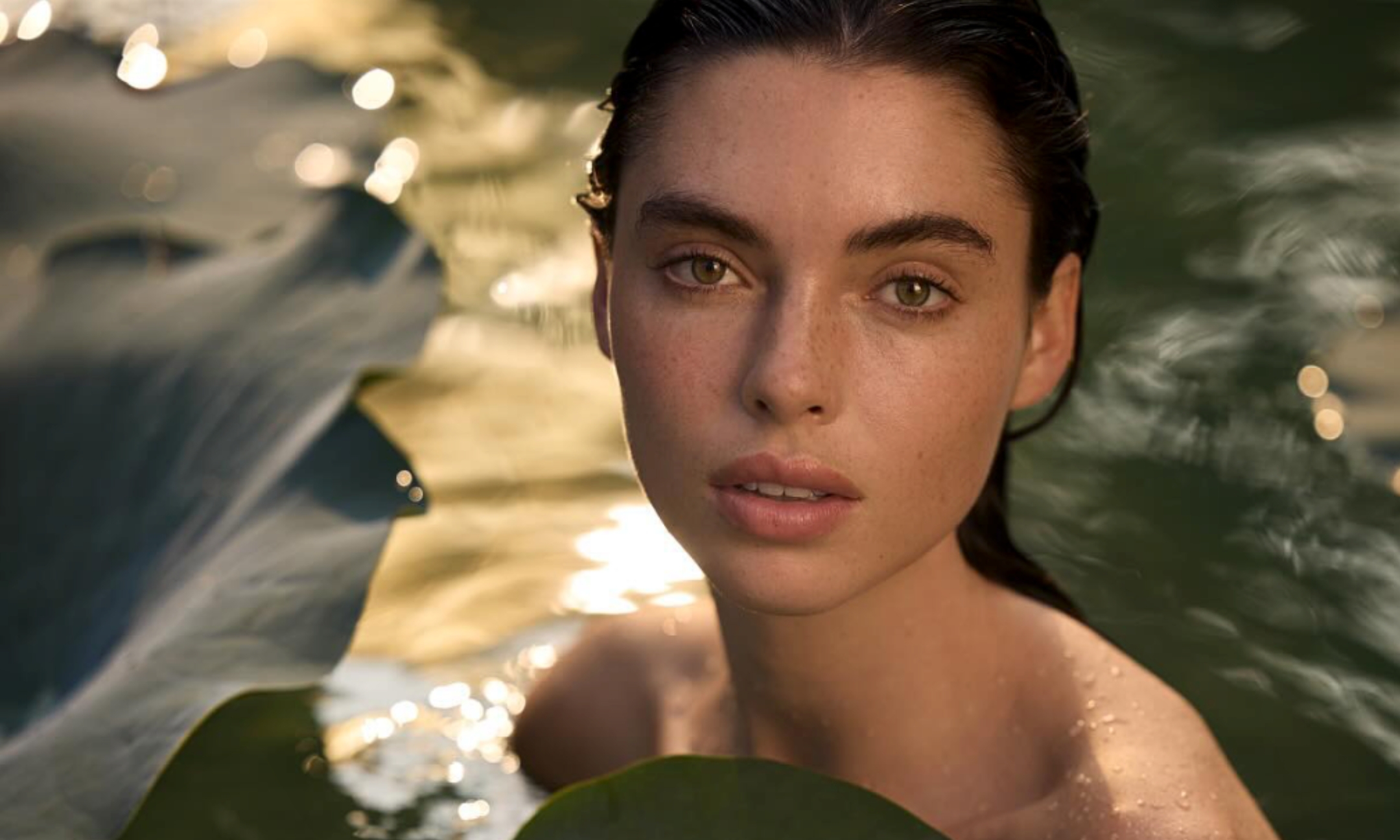A model with slicked-back hair and wet skin submerged in shimmering water next to large lily pads in a campaign image for Kama Ayurveda.