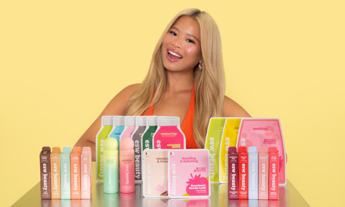 Beauty creator Katie Fang smiling behind a table filled with ESW Beauty skincare products, including sheet masks, eye patches, facial mists, and lip oils against a pastel yellow backdrop.
