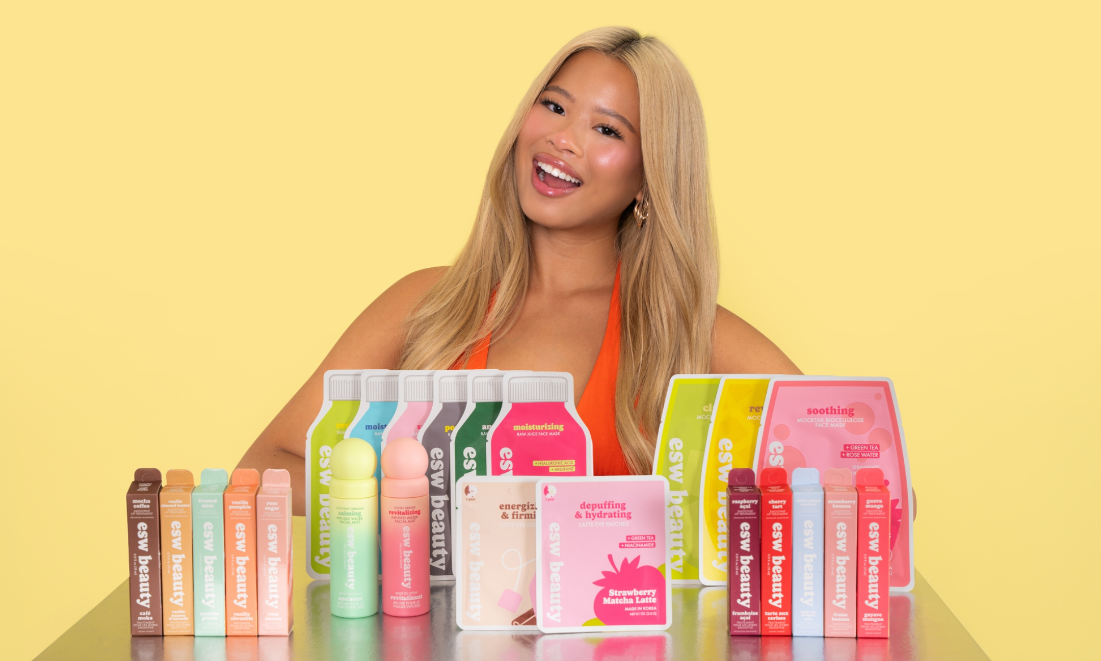 Beauty creator Katie Fang smiling behind a table filled with ESW Beauty skincare products, including sheet masks, eye patches, facial mists, and lip oils against a pastel yellow backdrop.