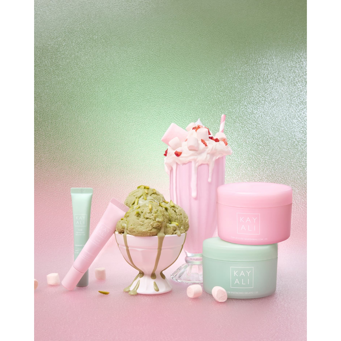 Kayali Yum Collection Cashmere Kiss Lip Balms and Silk Soufflé Body Creams arranged alongside scoops of pistachio ice cream and a pink marshmallow milkshake.