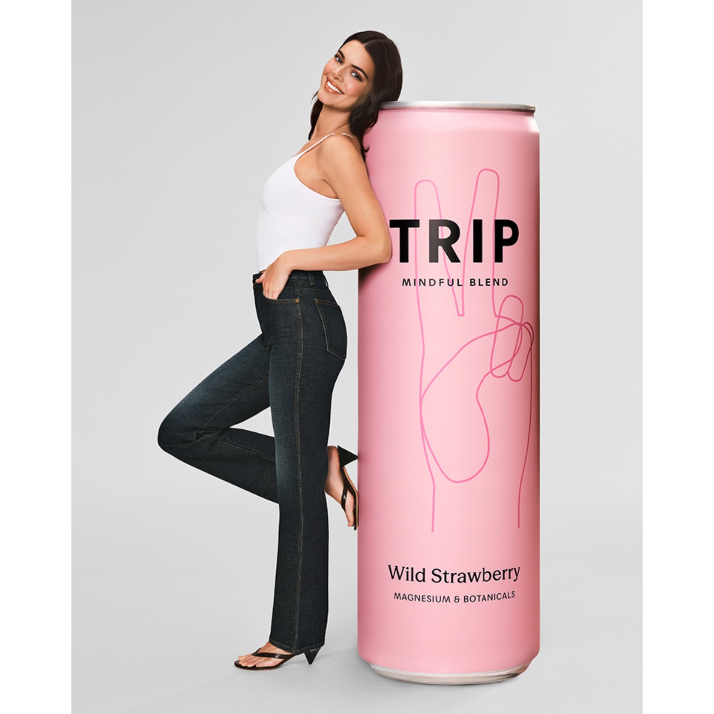 Kendall Jenner wearing a white tank top and dark denim jeans leaning against an oversized giant pink can of Trip Wild Strawberry drink against a plain gray studio backdrop.