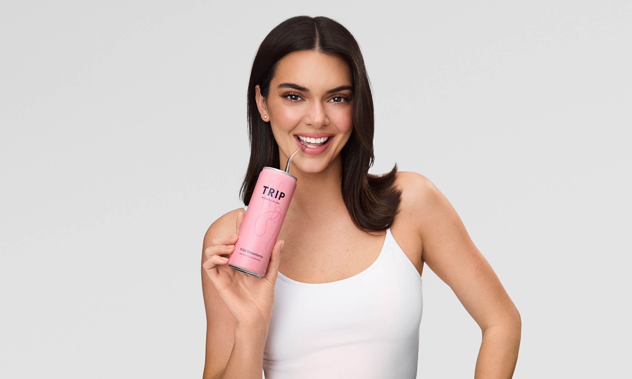 Kendall Jenner wearing a white tank top sipping through a straw from a pink can of Trip Wild Strawberry Mindful Blend drink against a light gray background.