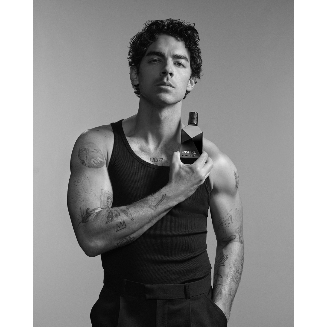 Black-and-white portrait of Joe Jonas in a dark tank top holding a bottle of Kenneth Cole Inspire fragrance near his shoulder.