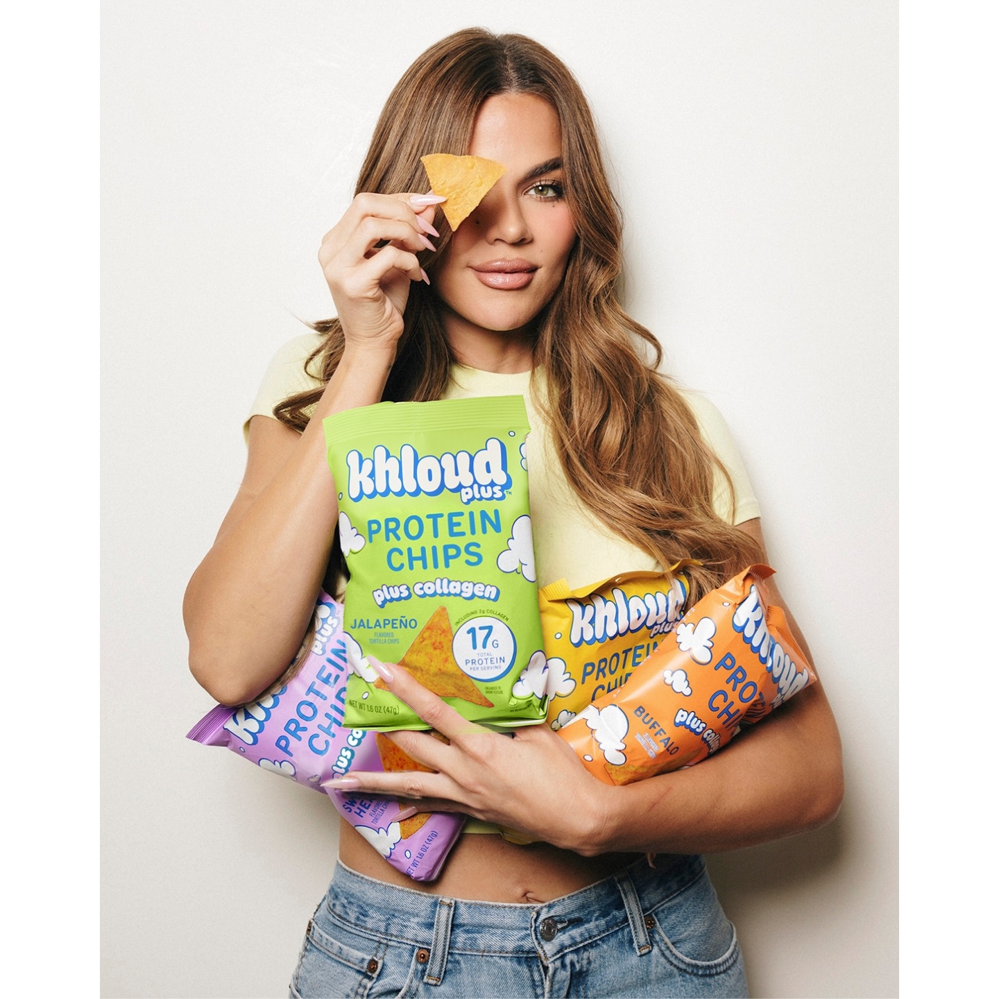 Khloé Kardashian posing with bags of Khloud Plus Protein Chips Plus Collagen, holding a tortilla chip over one eye against a white background.