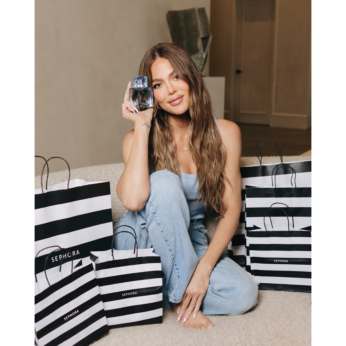 Khloé Kardashian sitting on a beige sofa surrounded by black-and-white striped Sephora shopping bags while holding a bottle of XO Blue Eau de Parfum.