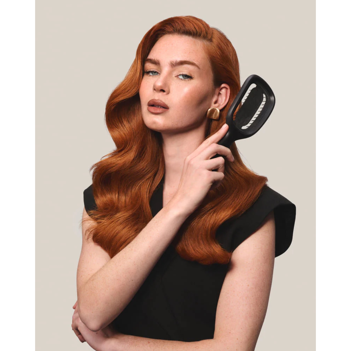 Model with wavy red hair in a black sleeveless outfit holding a black Killabrush hairbrush near her shoulder against a neutral background.