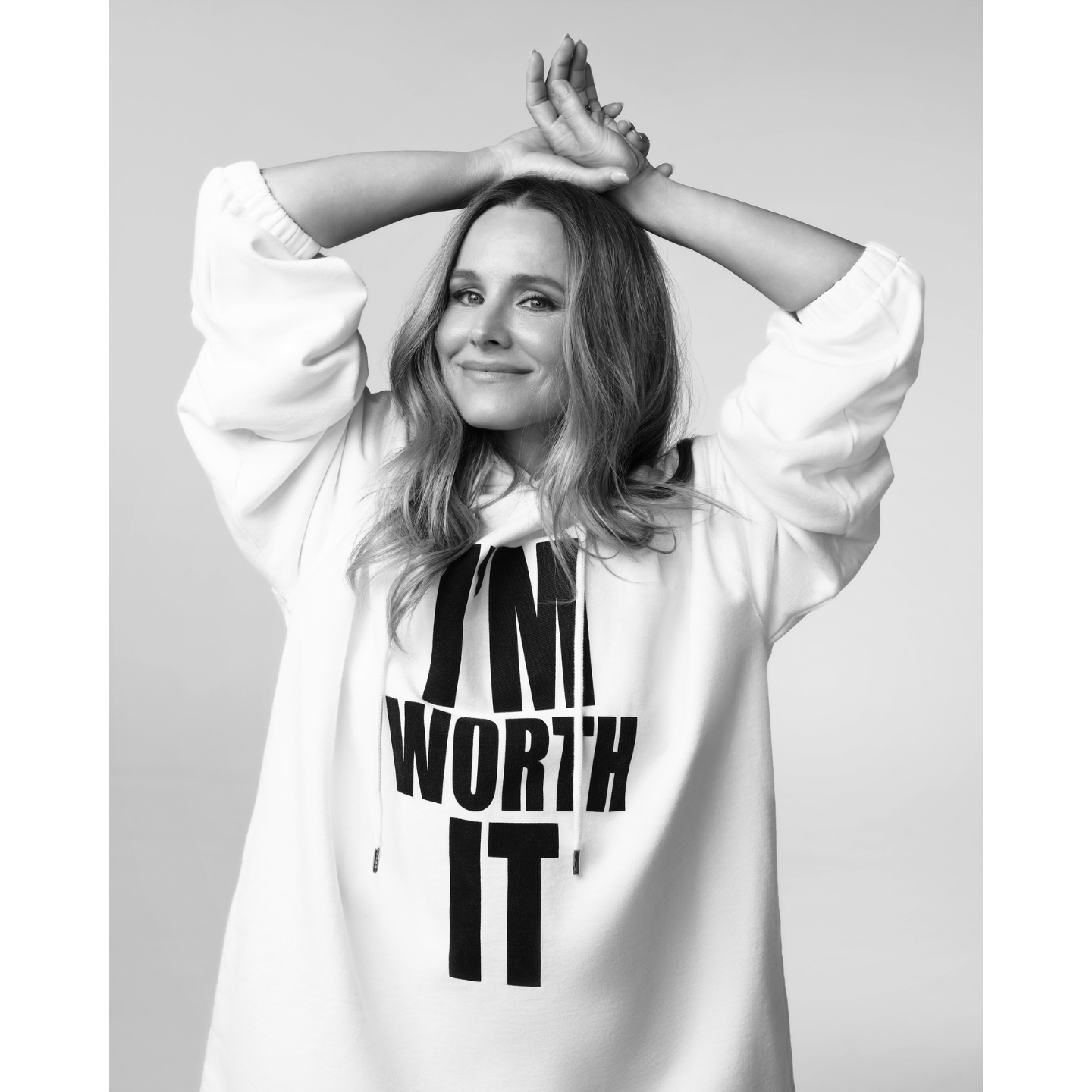 A black-and-white portrait of Kristen Bell posing with hands resting above her head wearing a white hoodie featuring L'Oréal's slogan "I'm Worth It".