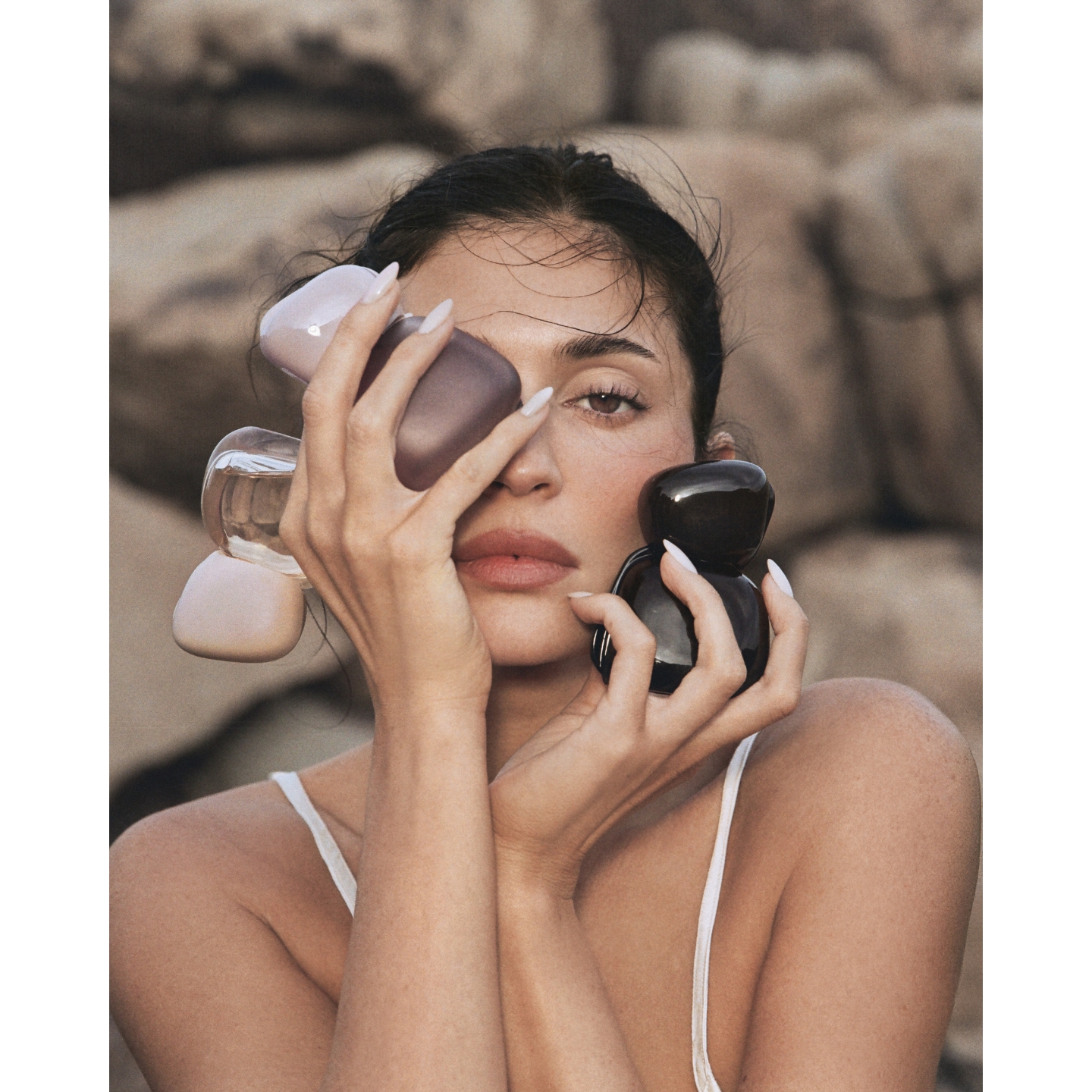 Kylie Jenner posing outdoors against a rocky stone backdrop, clutching the three pebble-shaped bottles of the Kylie Cosmetics Mood Stones fragrance collection to her face.