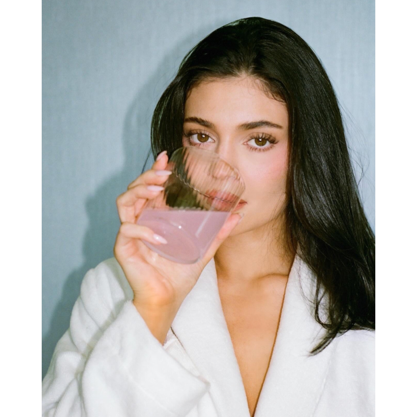 Kylie Jenner wearing a white bathrobe holding a glass of pink liquid up to her mouth for a k2o campaign image against a light blue background.
