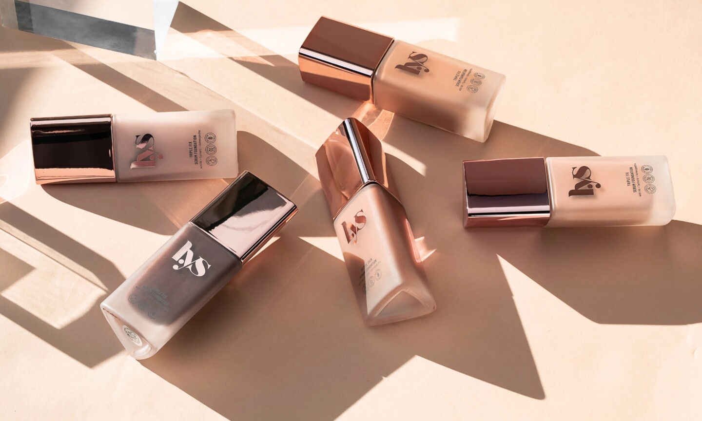 Five frosted glass bottles of LYS Beauty Triple Fix Serum Foundation lying flat on a beige surface with sharp geometric shadows, displaying a range of shades from fair to deep espresso.