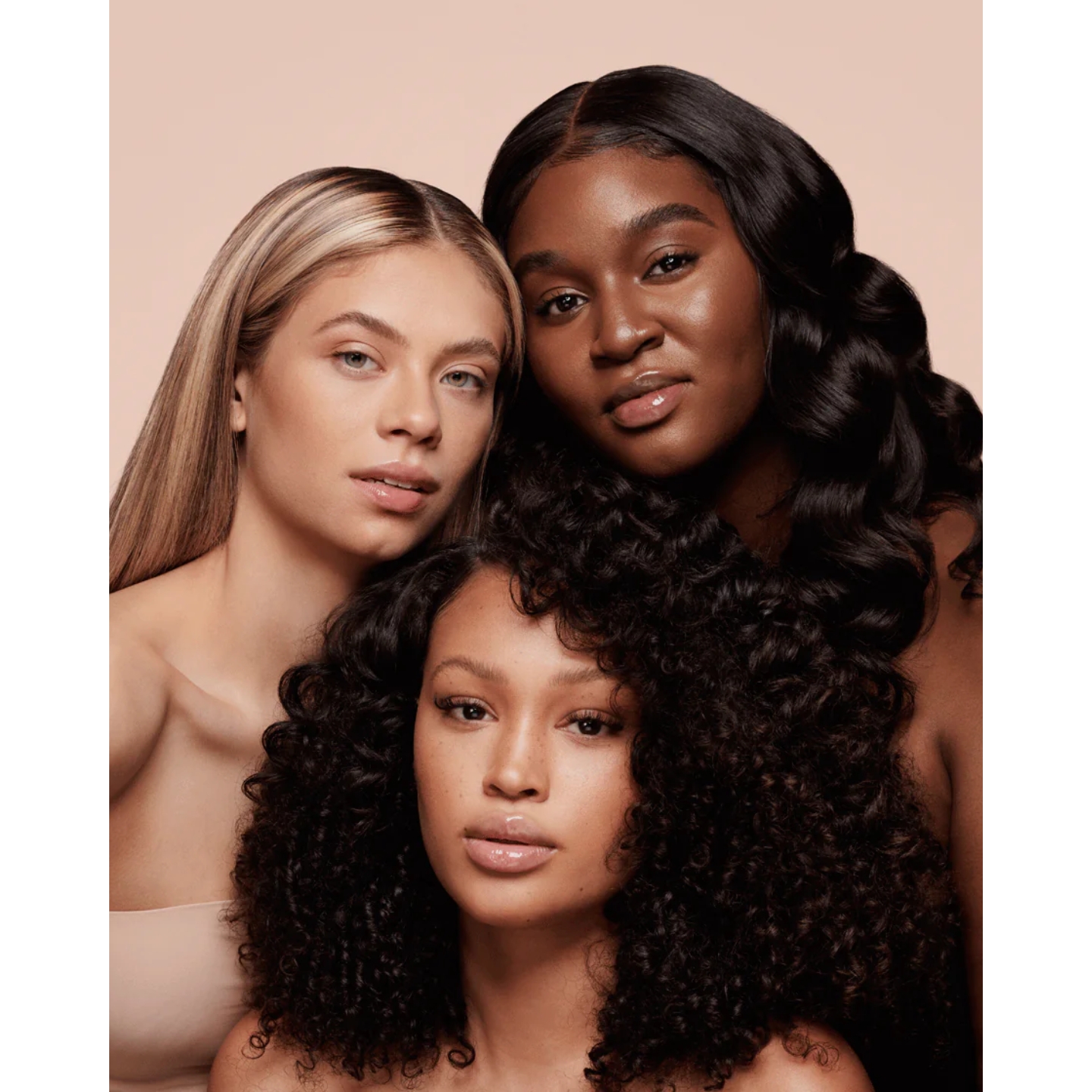 A close-up portrait of three models with diverse skin tones, hair textures, and radiant, natural complexions posing together against a soft beige background.