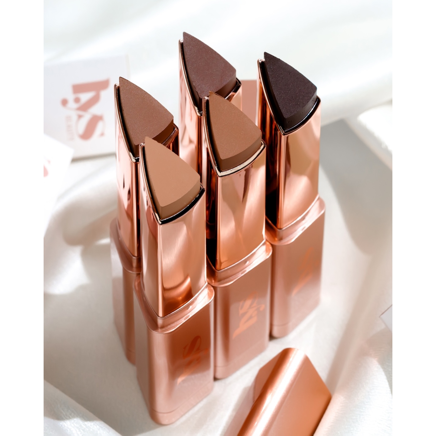 An open collection of LYS Beauty Triple Fix Skin Tint Foundation Sticks in rose gold triangular packaging, showcasing an inclusive range of shades from light tan to deep espresso.