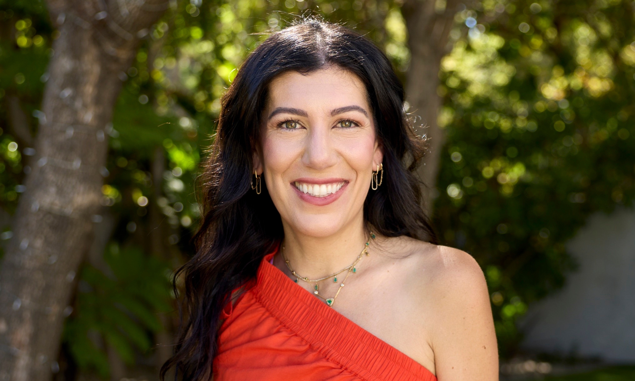 Portrait of Alleyoop founder Leila Kashani with long, wavy dark hair, wearing a red one-shoulder top and layered gold necklaces against a leafy green background.