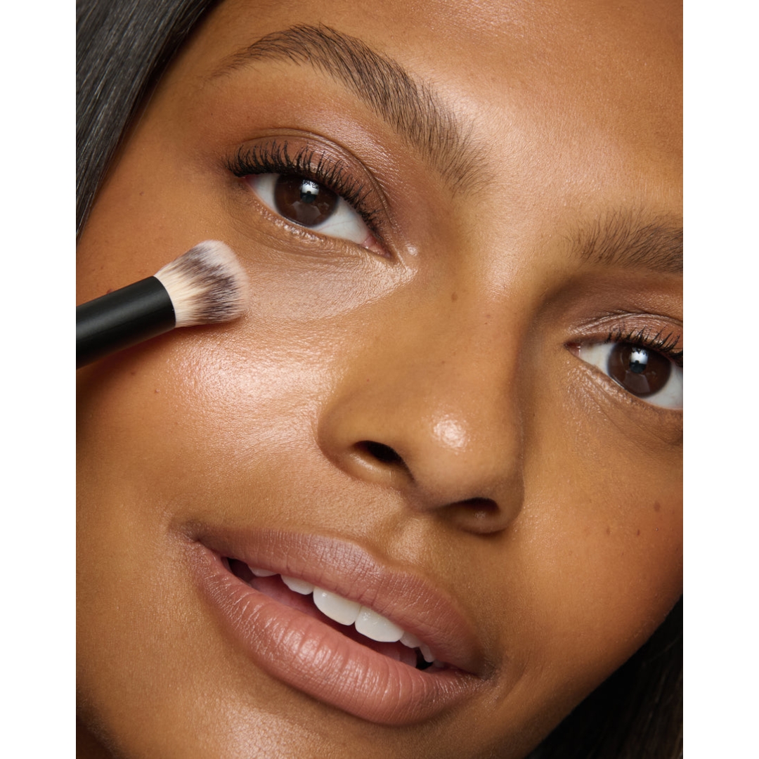 Extreme close-up portrait of a model applying Lisa Eldridge Luminist Concealer under her eye using a small fluffy makeup brush.