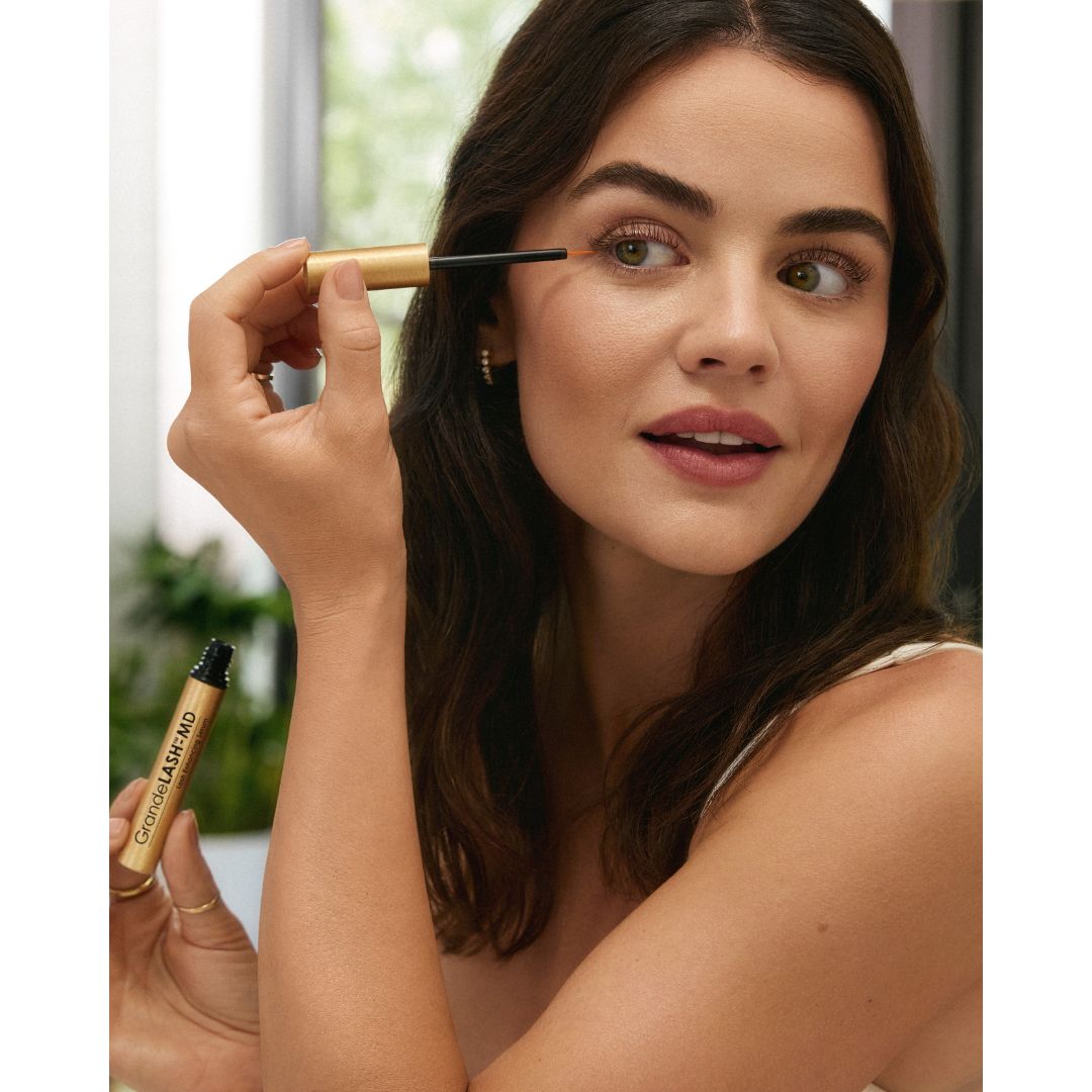 Lucy Hale applying GrandeLash-MD Lash Enhancing Serum to her upper lash line with a fine-tip applicator while holding the tube.