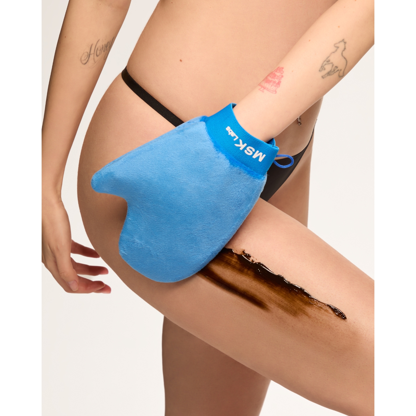A close-up of a model using a blue MSK Labs applicator mitt to apply self-tanner on her thigh, featured in the brand's Violet Grey US launch announcement on Beauty News Daily.