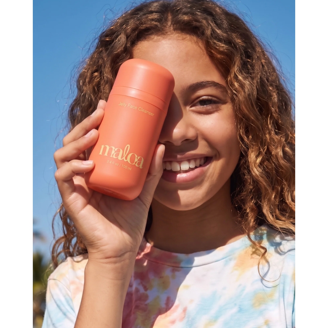 A smiling girl with long curly hair and a tie-dye T-shirt holding an orange bottle of Maloa Jelly Face Cleanser over one eye against a bright blue sky.