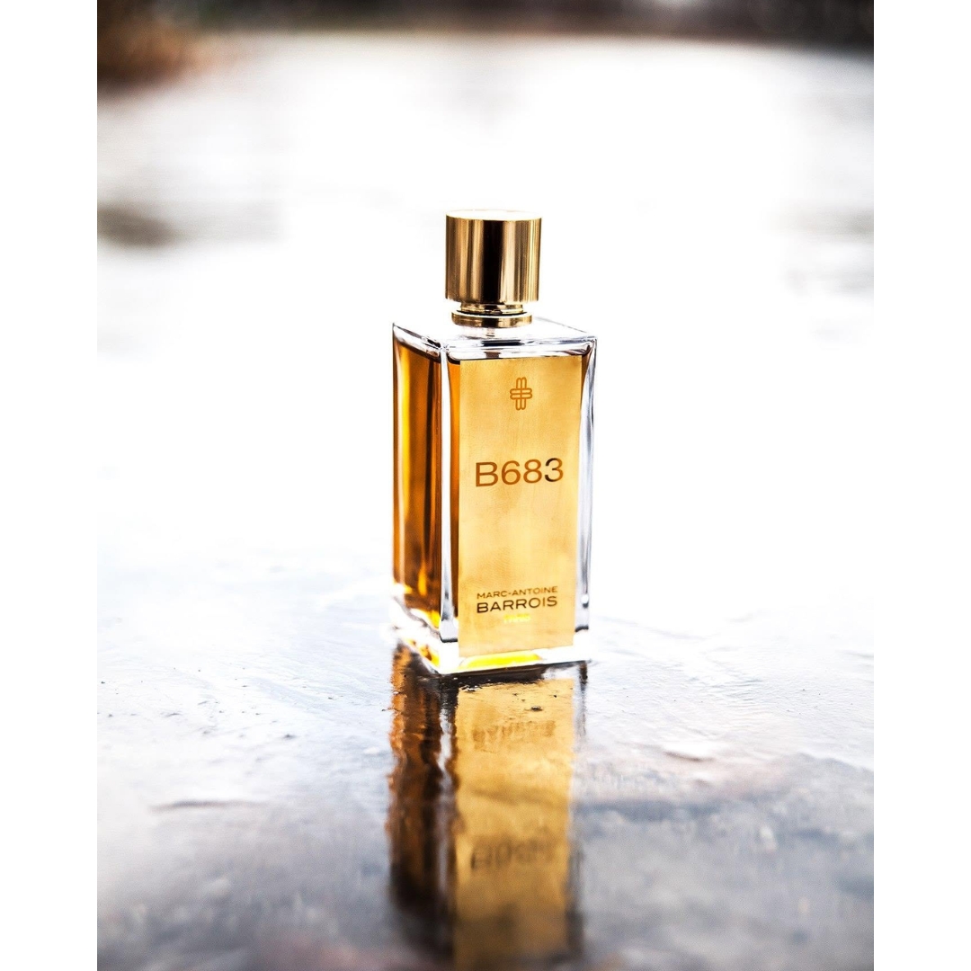 A glass bottle of Marc-Antoine Barrois B683 Eau de Parfum with a metallic gold label and cap standing upright on a reflective wet surface outdoors.