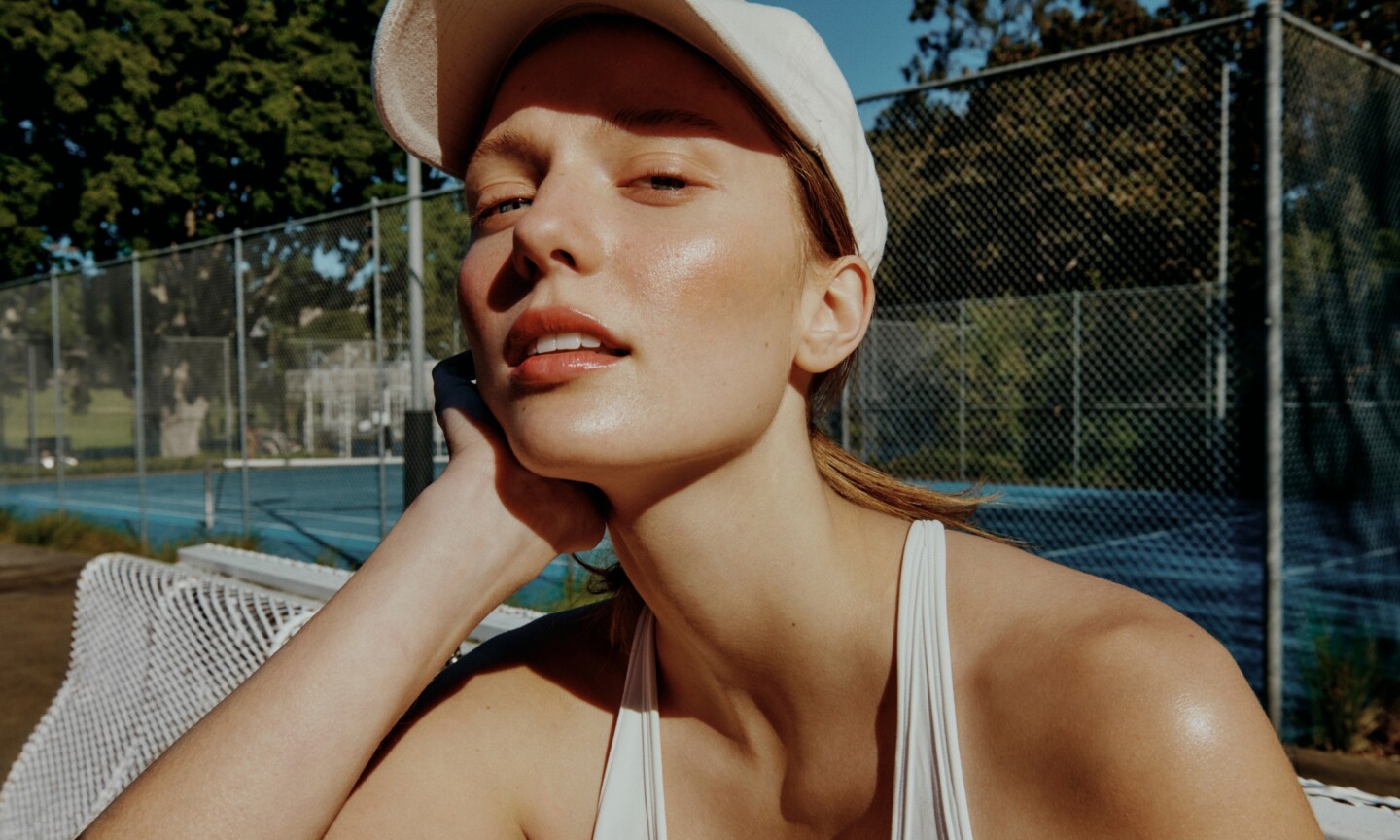 Model with glowing, sun-protected skin wearing a white cap on a tennis court, featured in the Beauty News Daily best facial SPFs and sunscreens roundup.