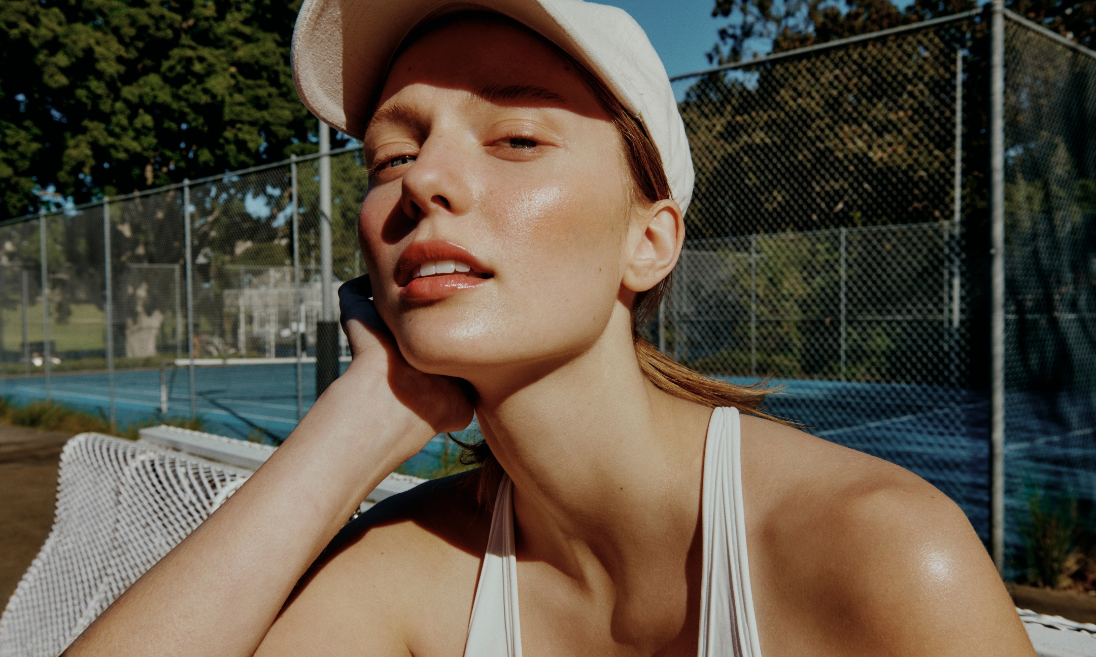 Model with glowing, sun-protected skin wearing a white cap on a tennis court, featured in the Beauty News Daily best facial SPFs and sunscreens roundup.