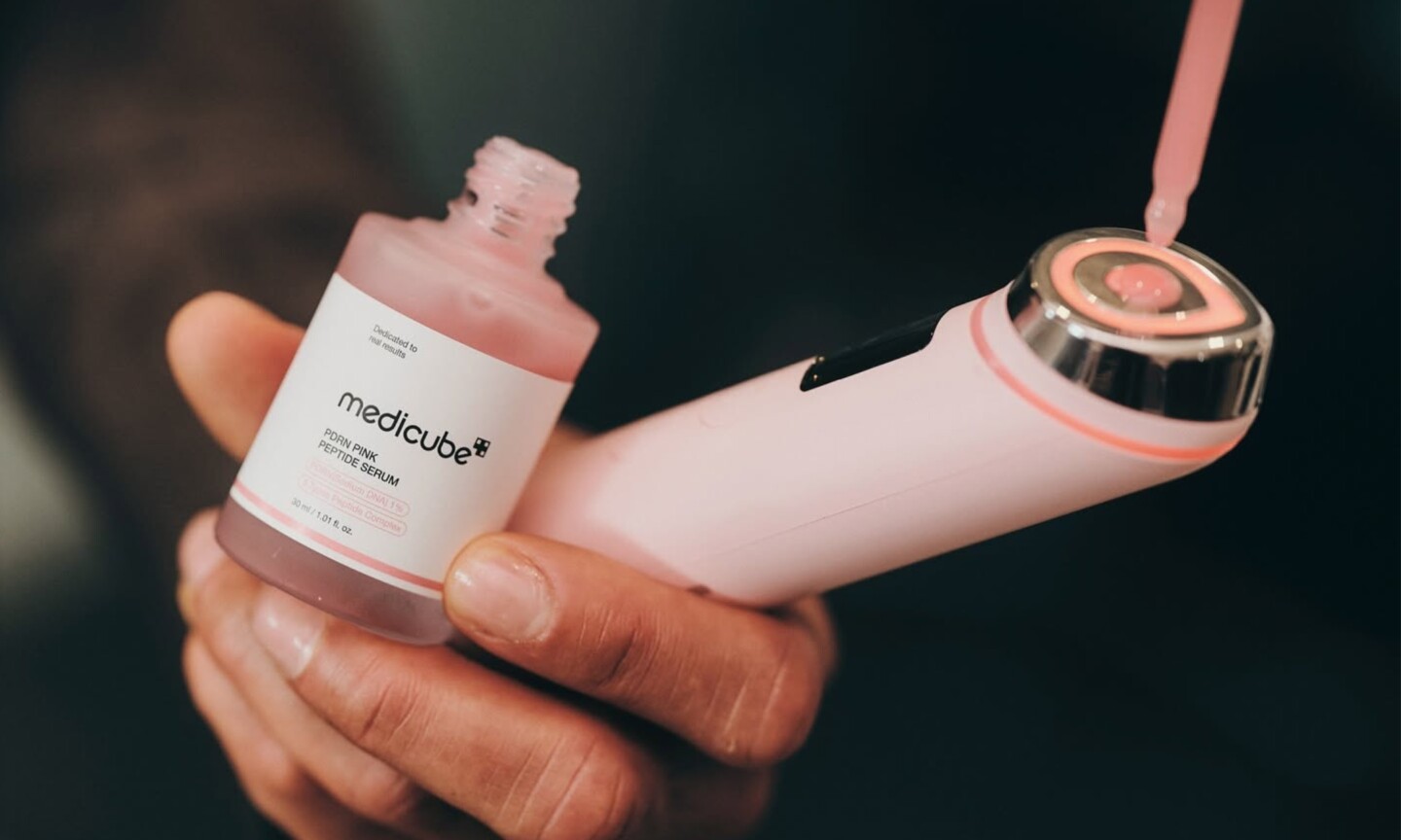 A hand holding Medicube PDRN Pink Peptide Serum and the pink Booster Pro X2 as a dropper applies serum to the device head