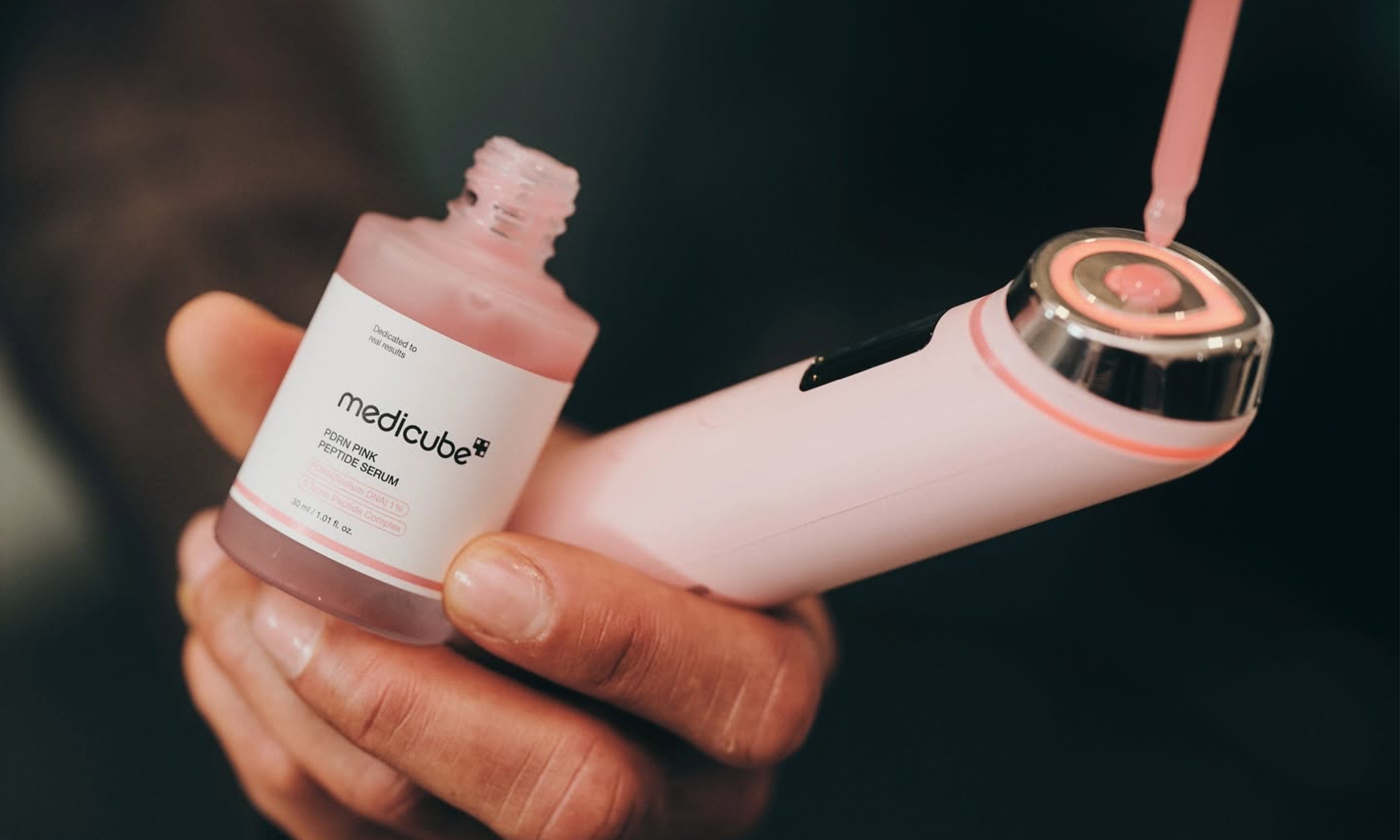 A hand holding Medicube PDRN Pink Peptide Serum and the pink Booster Pro X2 as a dropper applies serum to the device head