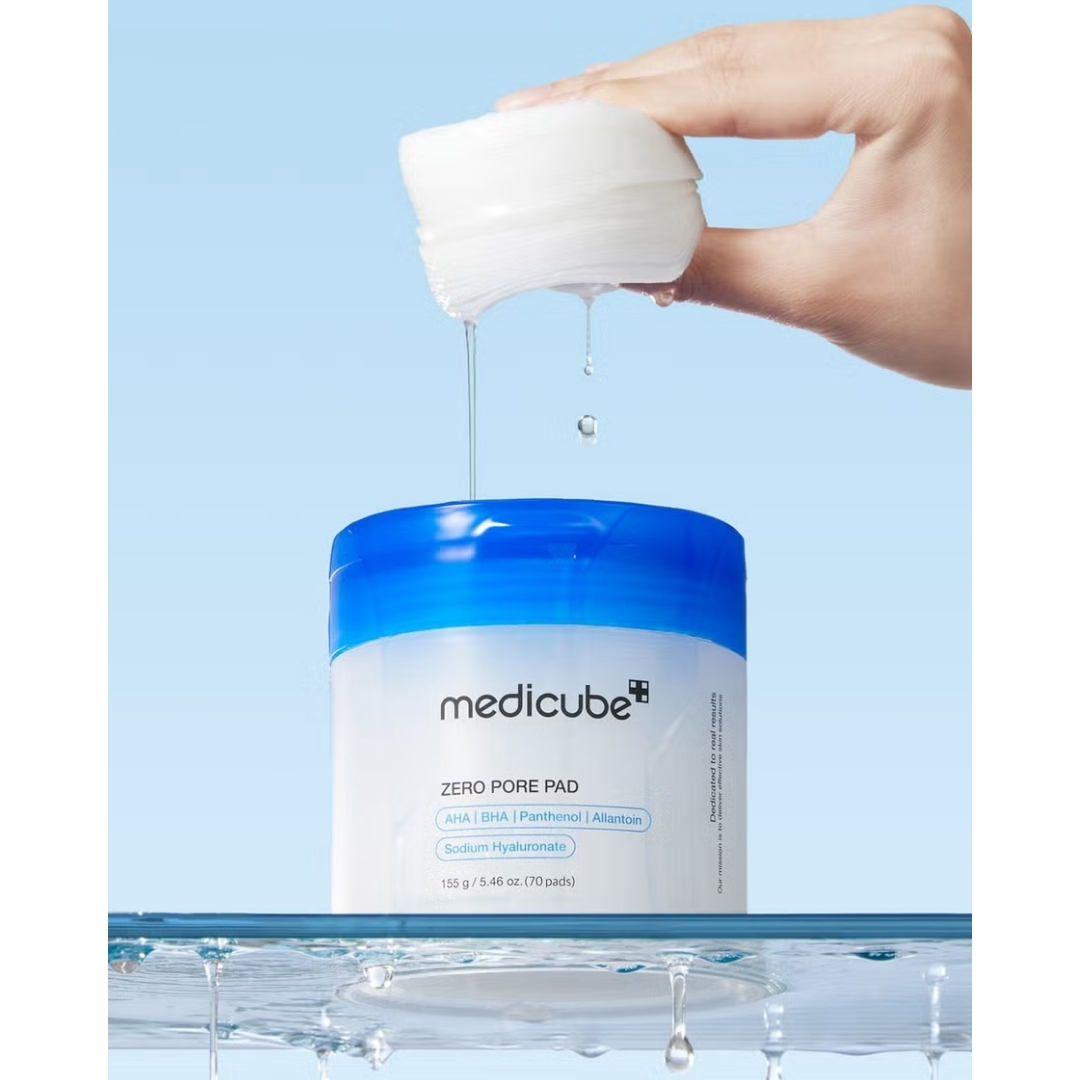 A hand squeezing a stack of saturated toner pads over an open white and blue jar of Medicube Zero Pore Pads, dripping serum into the container.