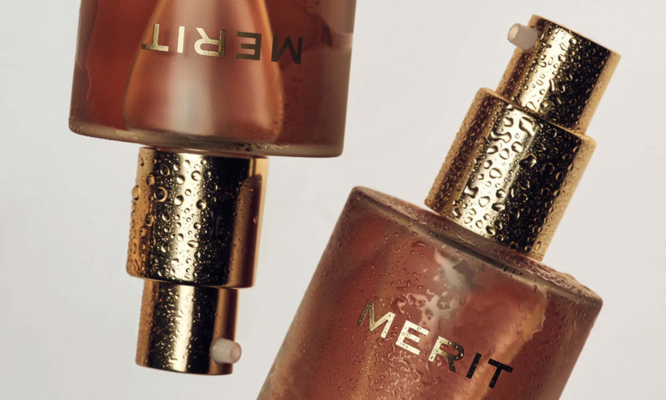 Two glass bottles of Merit Great Skin Instant Glow Serum with gold pump dispensers covered in fine water droplets, set against an off-white background.
