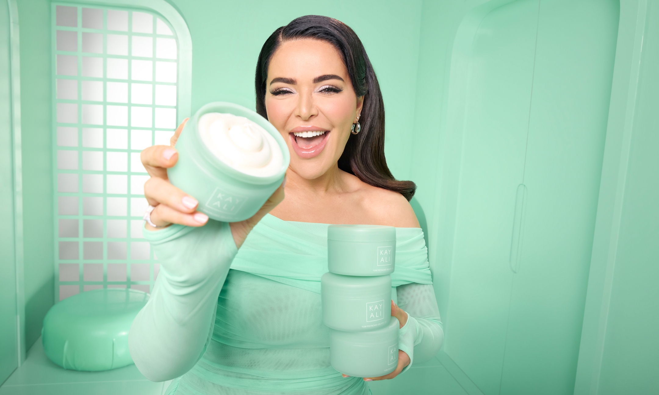 Kayali founder Mona Kattan smiling in a pastel green room while holding an open tub of Kayali Yum Pistachio Gelato Silk Soufflé Body Cream.