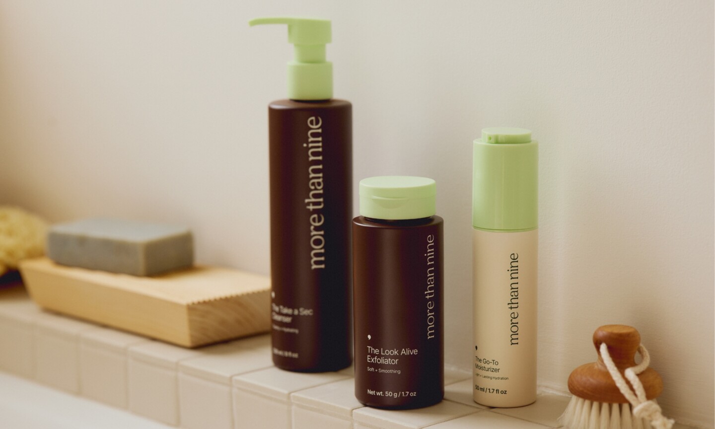 Three More Than Nine skincare products arranged on a tiled bathroom ledge, featuring the Take A Sec Cleanser and The Look Alive Exfoliator in dark brown bottles alongside The Go-To Moisturizer in a beige bottle with light green caps.