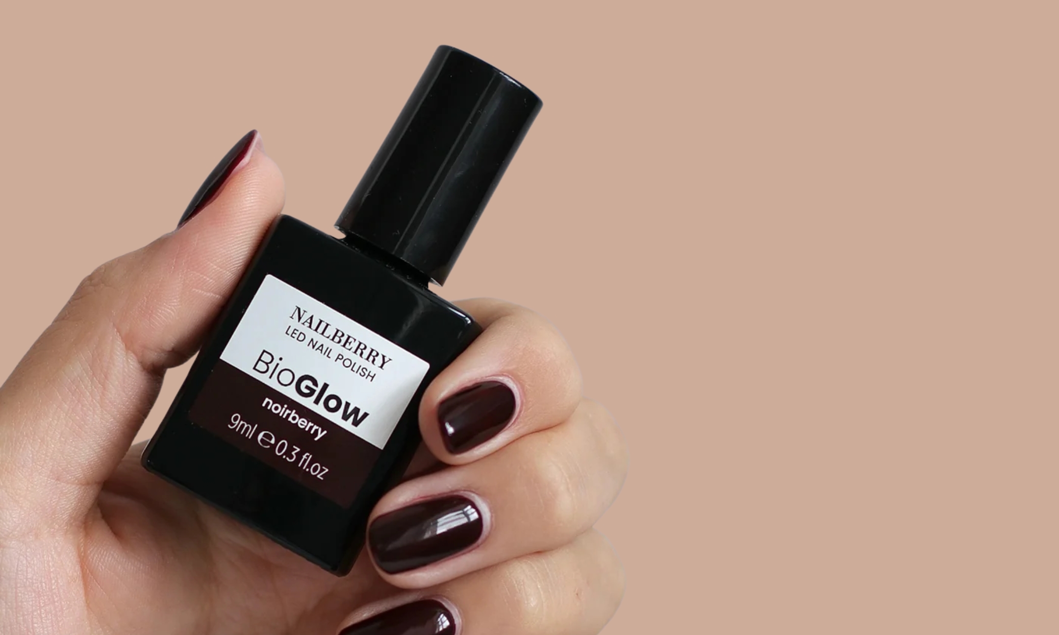 A hand with deep dark berry manicured nails holding a bottle of Nailberry BioGlow LED Nail Polish in the shade Noirberry against a beige background.