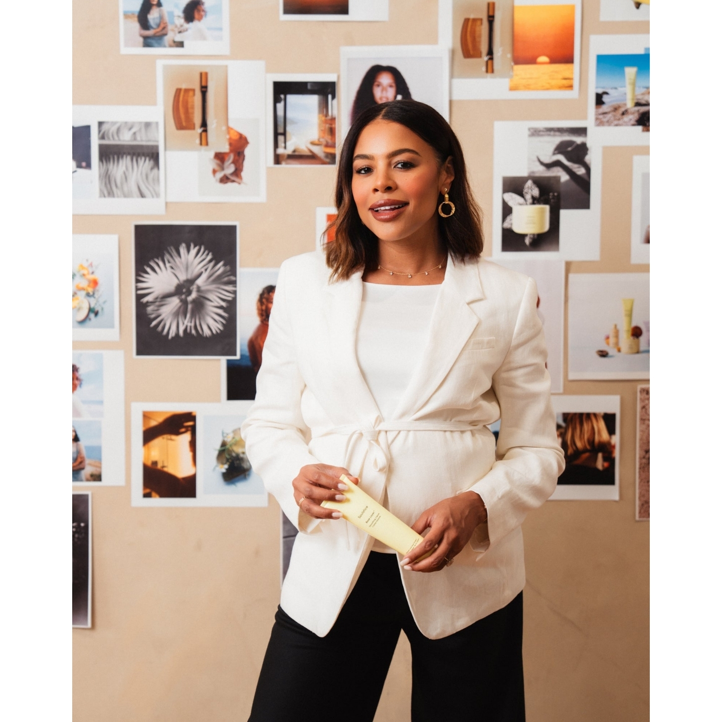 Nancy Twine, founder of Soulstice, wearing a white blazer while holding the Soulstice Base Layer Smoothing Cream in front of a photo mood board.