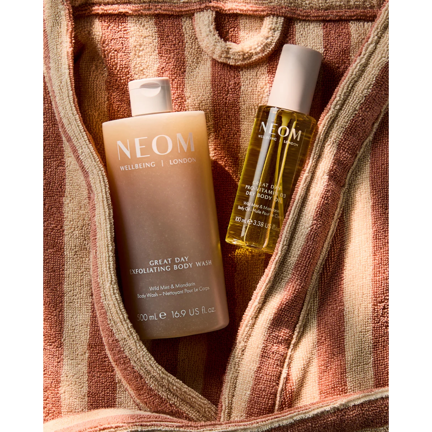 Neom Wellbeing Great Day Exfoliating Body Wash and Great Day Pro-Vitamin D3 Dry Body Oil in Wild Mint and Mandarin resting inside the collar of a brown and beige striped bathrobe.