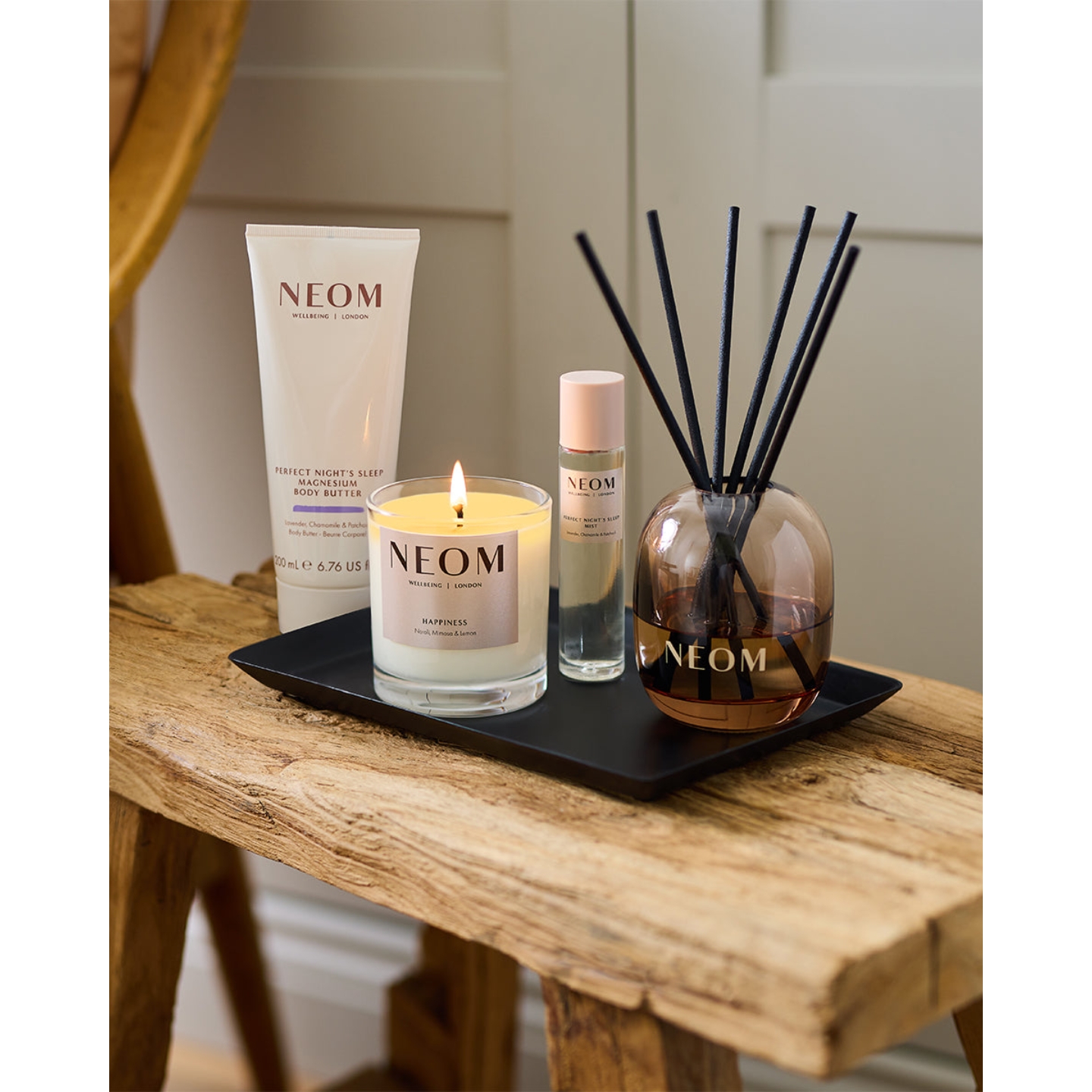 A Neom Wellbeing set arranged on a rustic wooden bench, including a lit Happiness scented candle, a glass reed diffuser, Perfect Night's Sleep mist, and Perfect Night's Sleep Magnesium Body Butter.