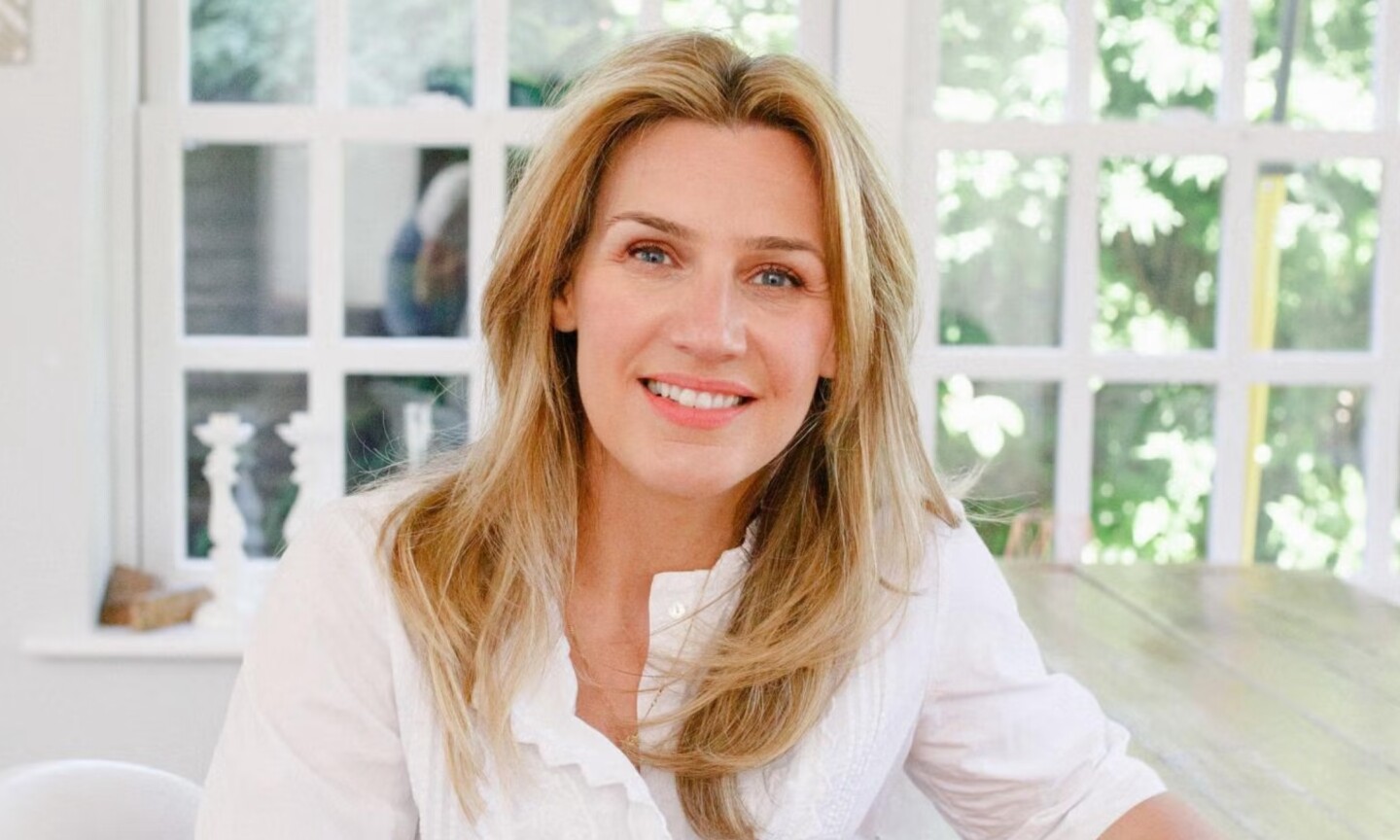 Portrait of Neom Wellbeing founder Nicola Elliott wearing a white button-up top, smiling naturally in front of a bright window background.