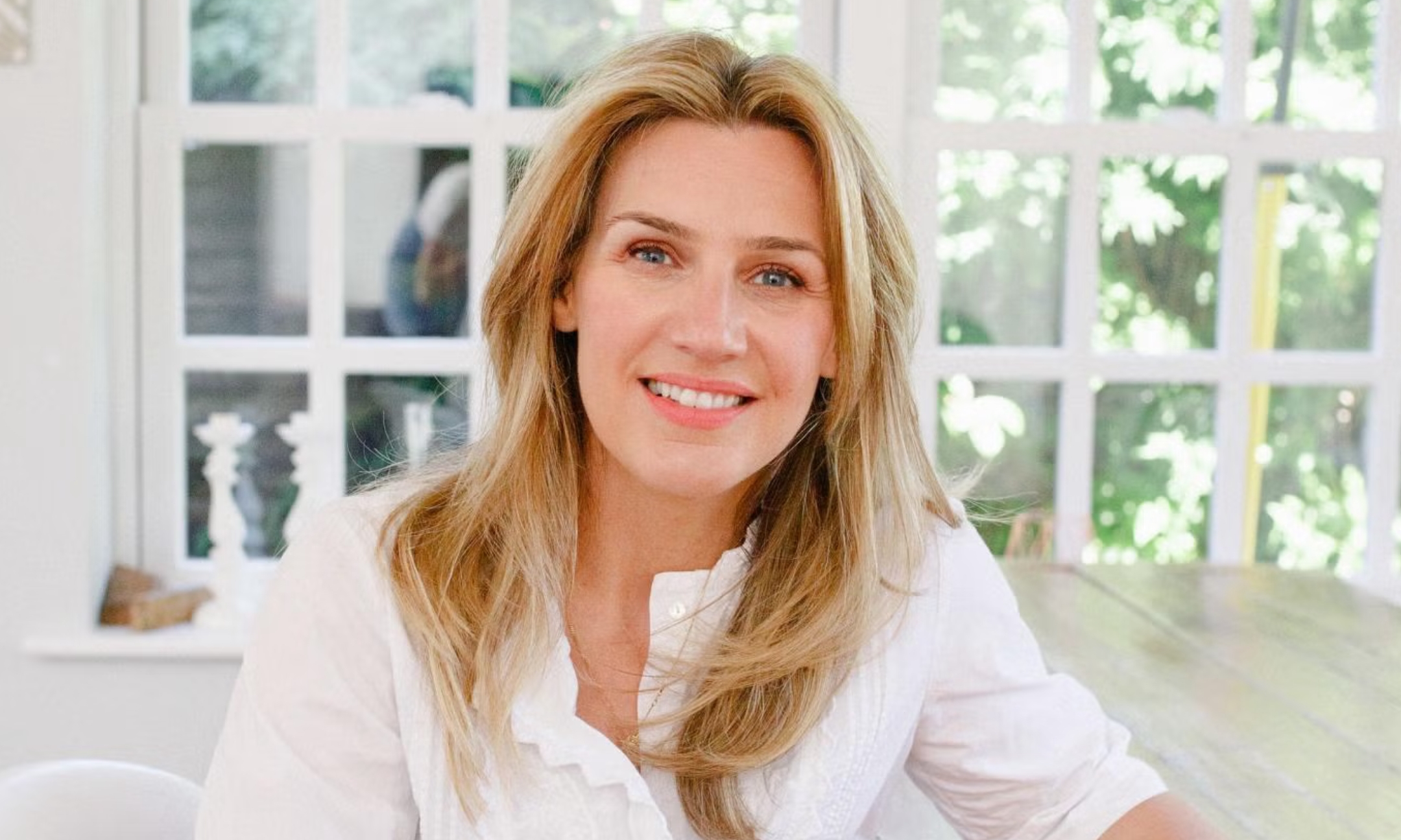 Portrait of Neom Wellbeing founder Nicola Elliott wearing a white button-up top, smiling naturally in front of a bright window background.