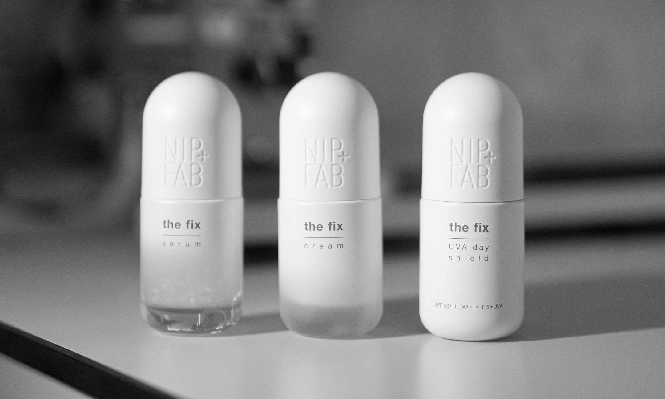 A black-and-white studio shot of three Nip+Fab The Fix products in rounded white pill-like bottles: Serum, Cream, and UVA Day Shield.