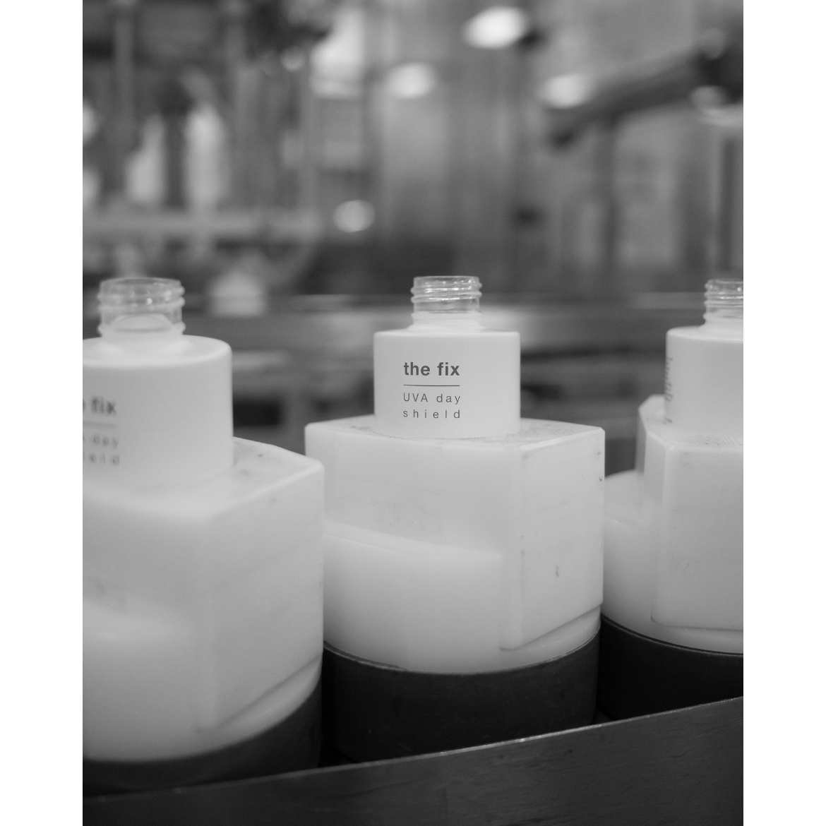 A black-and-white close-up of uncapped Nip+Fab The Fix UVA Day Shield bottles sitting in holders on a factory production line.
