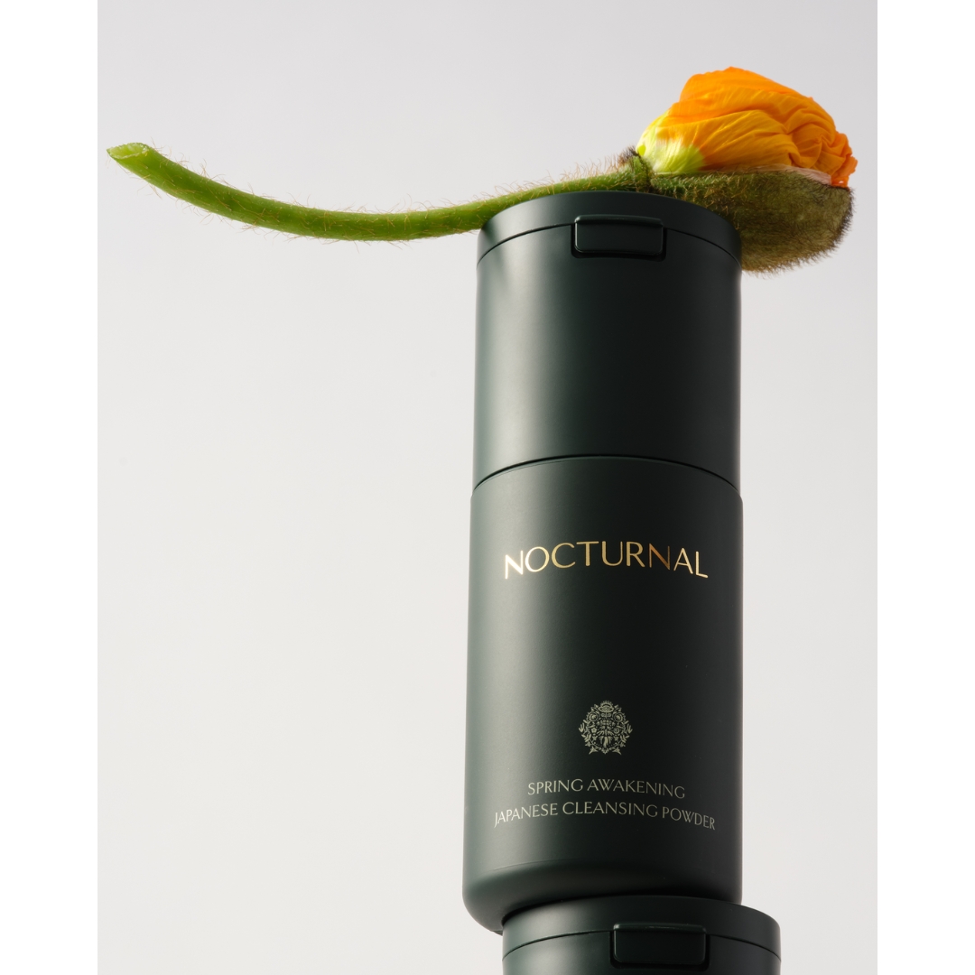 A cylindrical dark green bottle of Nocturnal Spring Awakening Japanese Cleansing Powder stacked on top of another bottle, balanced with an orange poppy bud lying across the top against a light background.