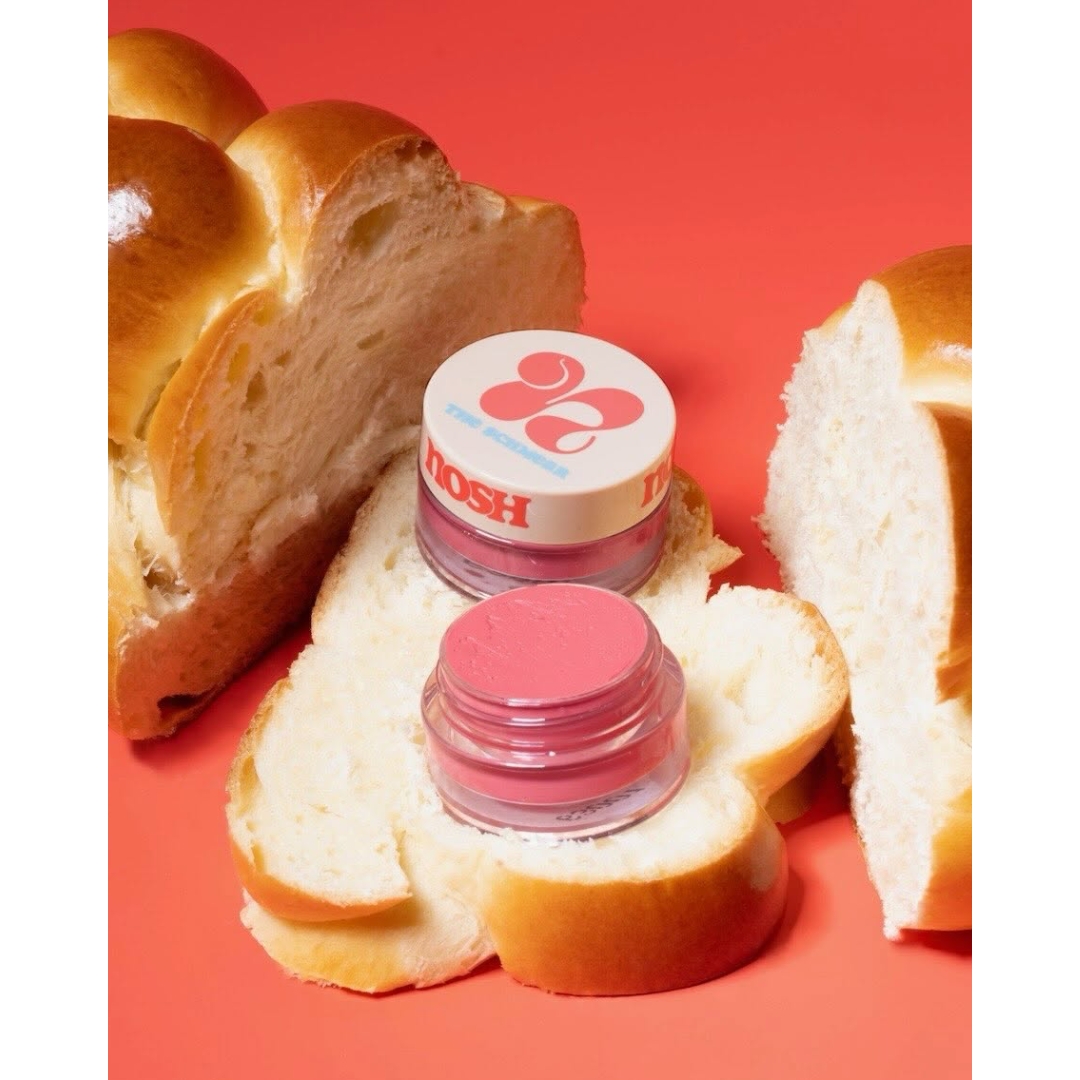 Two small glass pots of pink Nosh Beauty The Schmear lip product—one open and one capped—resting on slices of braided challah bread against an orange background.