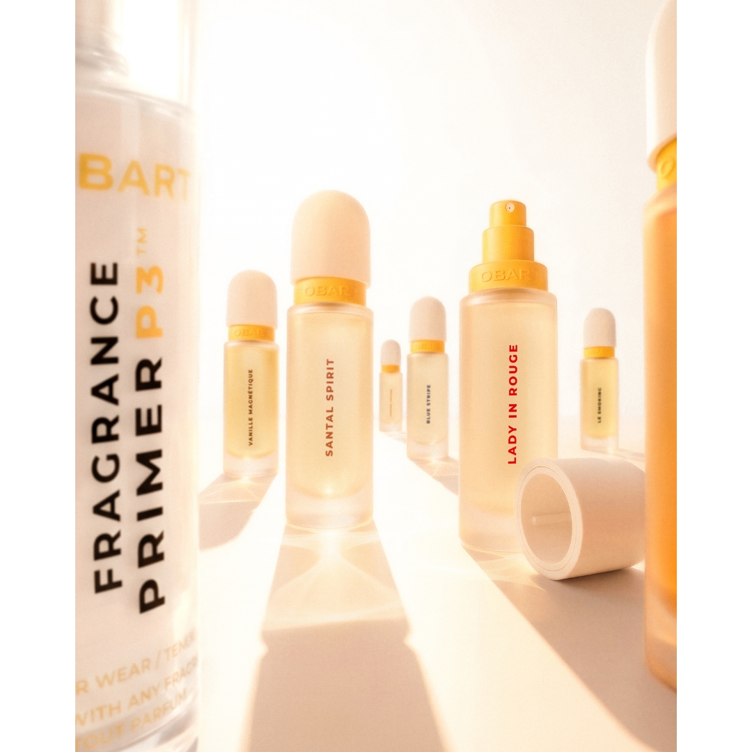 An array of frosted glass Obart perfume bottles, including Santal Spirit and Lady in Rouge, displayed alongside the Obart Fragrance Primer P3 in warm, bright sunlight.