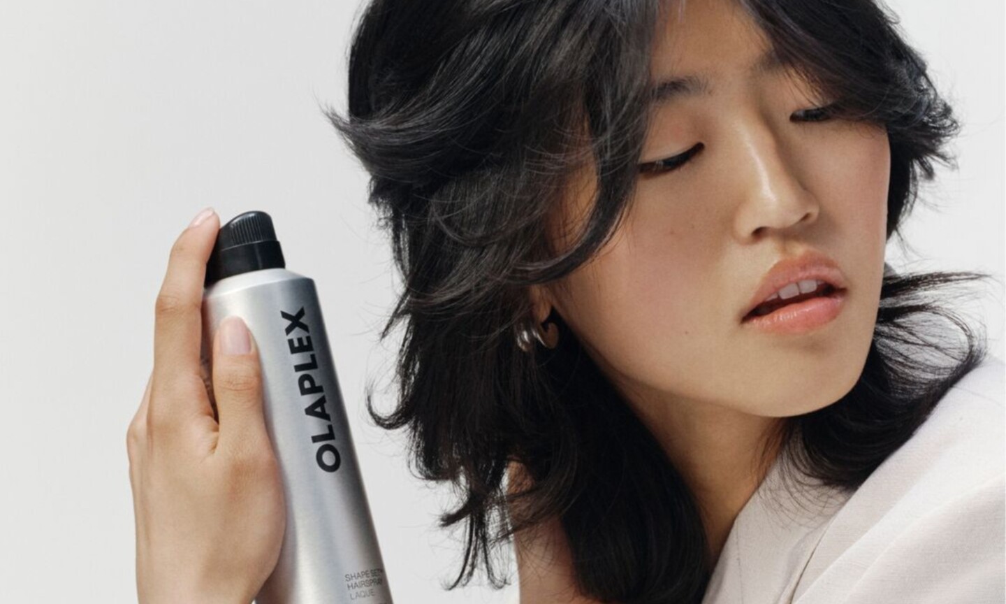 Close-up of a model with short black layered hair holding a silver can of Olaplex Shape Set Hairspray near her face against a plain light gray background.