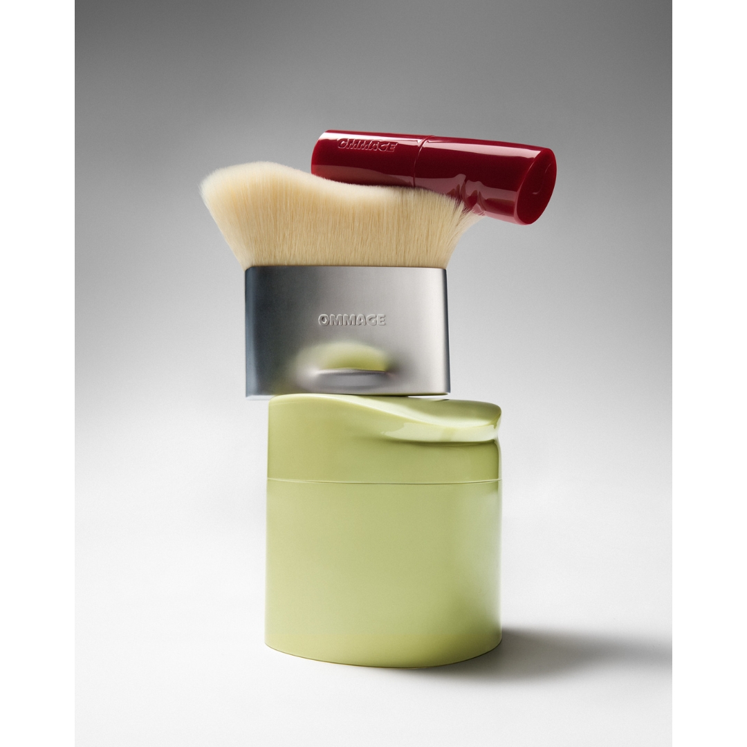 A vertical product stack featuring the Ommage First Base, Buffer Brush, and Lip Coat resting on top.