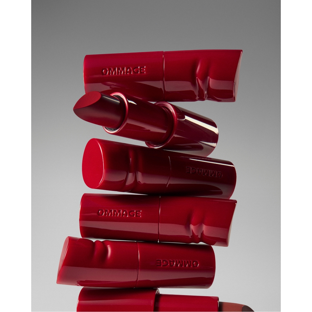 A vertical arrangement of stacked red Ommage Lip Coat tubes with one uncapped bullet revealed near the top against a grey studio background.