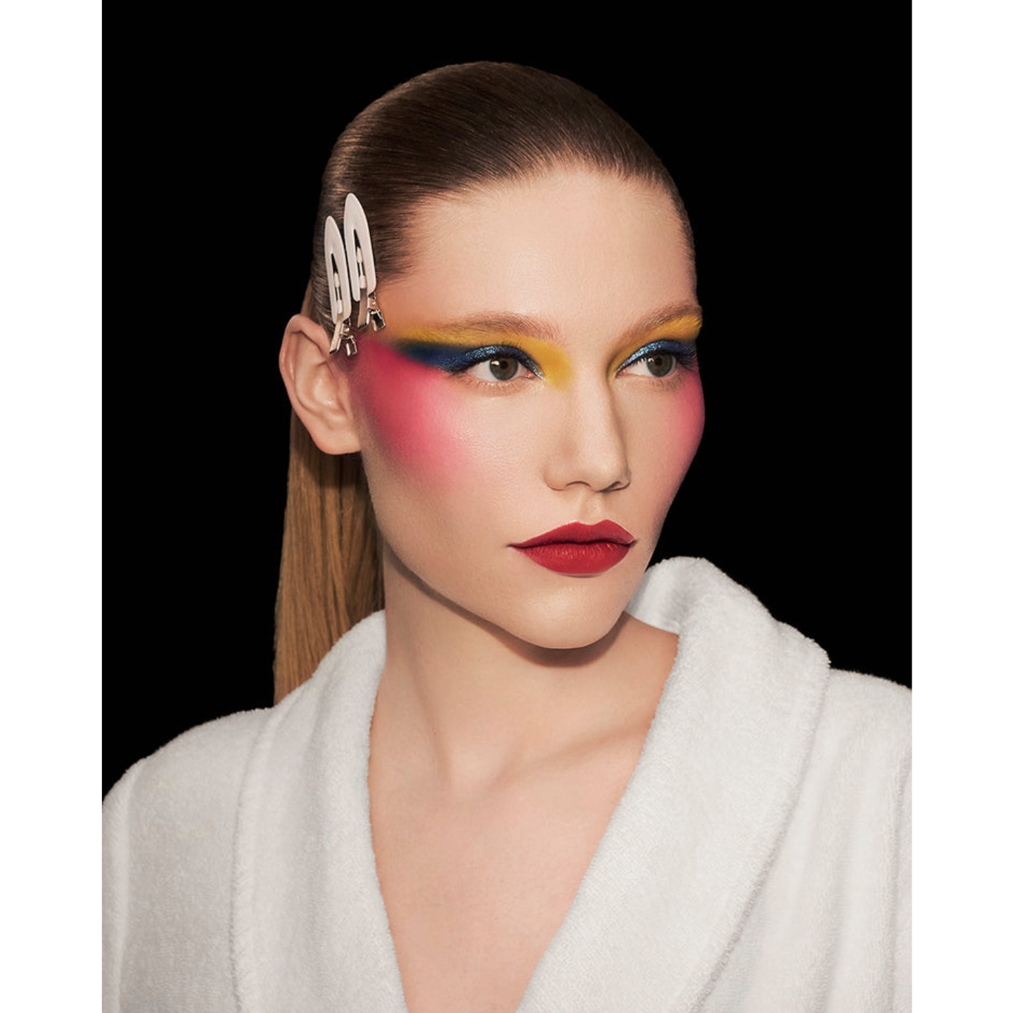 Model in a white plush bathrobe with hair styled in flat clips wearing vibrant yellow, shimmer blue, and bright pink eyeshadow paired with bold red lipstick in a campaign image for the Pat McGrath Labs Backstage Artistry Collection.
