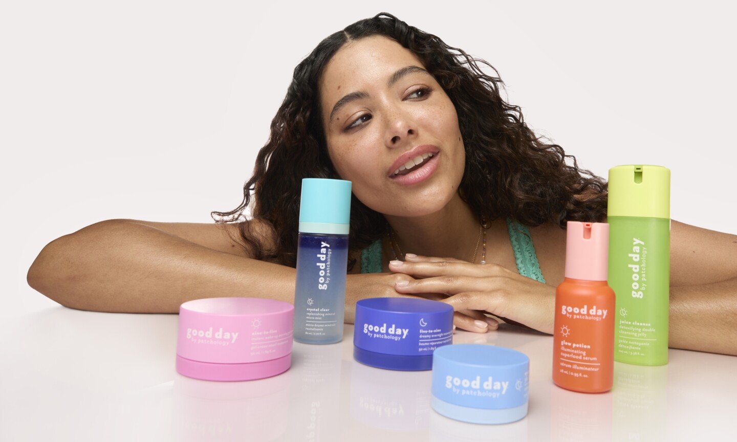 A model with dark curly hair leaning over a desk showcasing the full Good Day by Patchology skincare range, featuring colorful tubs, serums, mists, and cleansers.