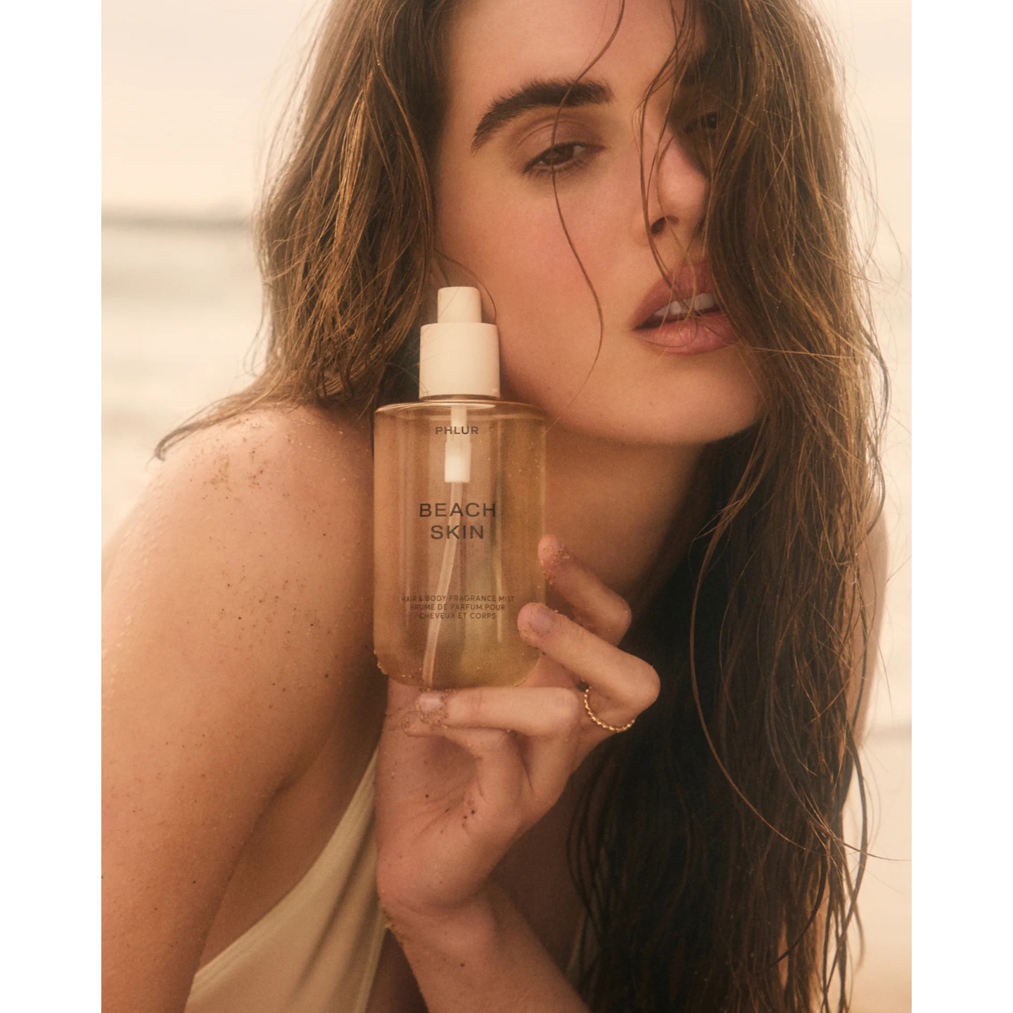 Model with wet hair and sun-kissed skin holding a bottle of Phlur Beach Skin Hair & Body Fragrance Mist near her face at the beach
