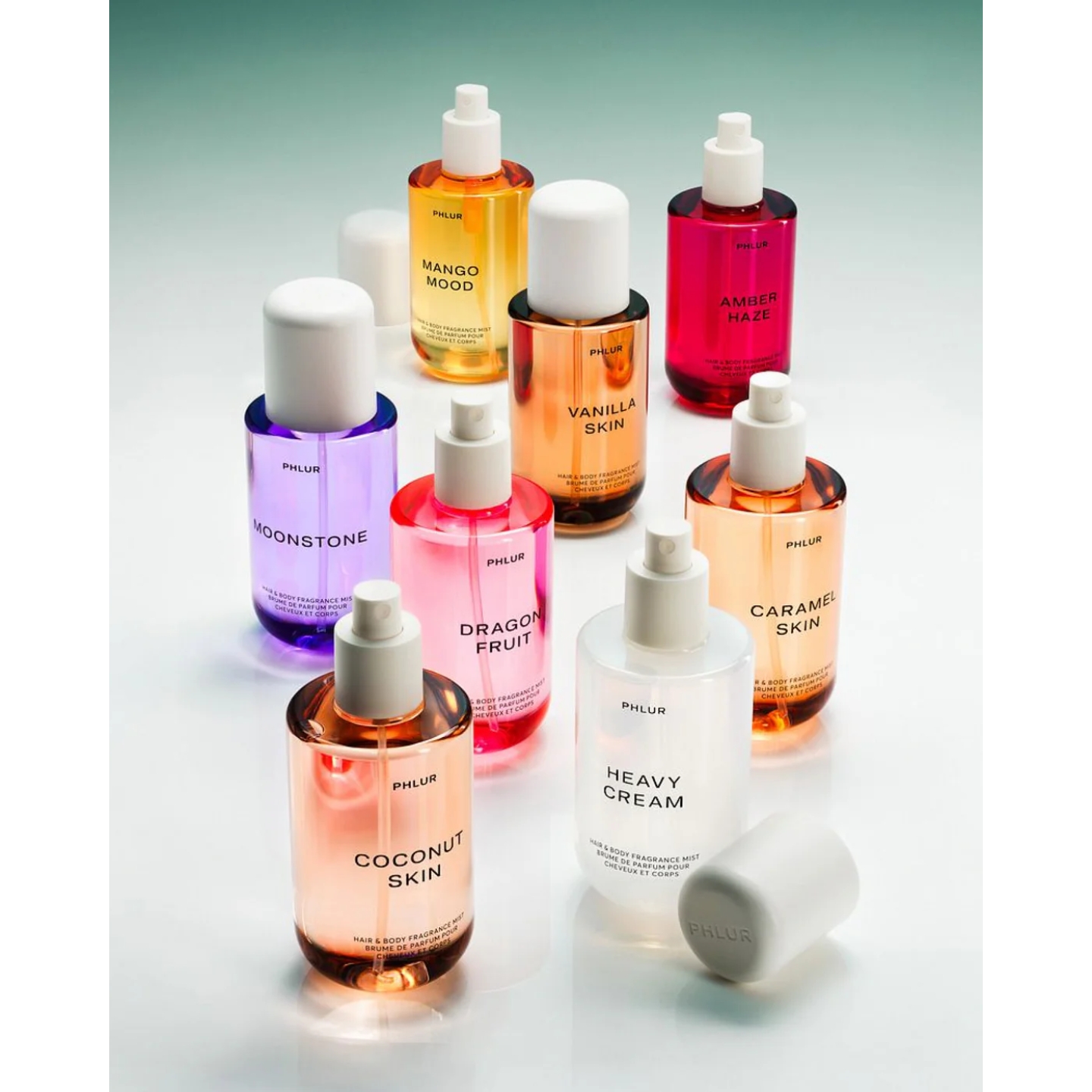 An assortment of Phlur Hair & Body Fragrance Mists displayed on a reflective surface, including Coconut Skin, Heavy Cream, Dragon Fruit, Moonstone, Vanilla Skin, Caramel Skin, Mango Mood, and Amber Haze.